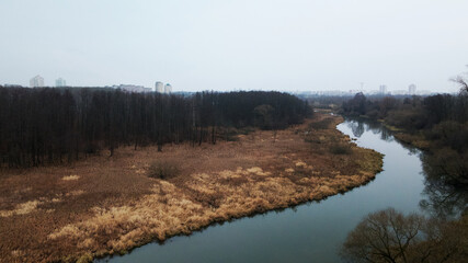 The river among the city park. Late autumn, cloudy. Trees without leaves and dried reeds along the banks of the river. The city is visible on the horizon. Aerial photography.