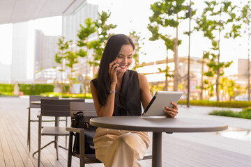 Portrait of beautiful Asian woman sitting outdoors at coffee shop restaurant talking on phone and using digital tablet computer