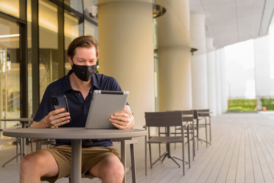 Man Sitting Outdoors At Coffee Shop Restaurant Social Distancing And Wearing Face Mask To Protect From Covid 19 While Using Phone And Digital Tablet