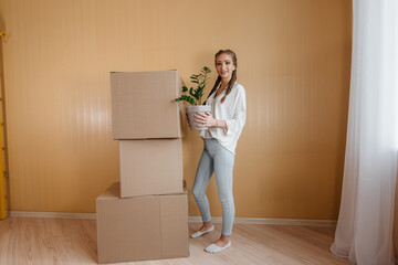 A young woman stands with a houseplant after moving to a new apartment against the background of unpacked things. Purchase of real estate. Housewarming, delivery and transportation of goods.