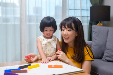 Young mom helping daughter drawing with colored pencils in living room at home.