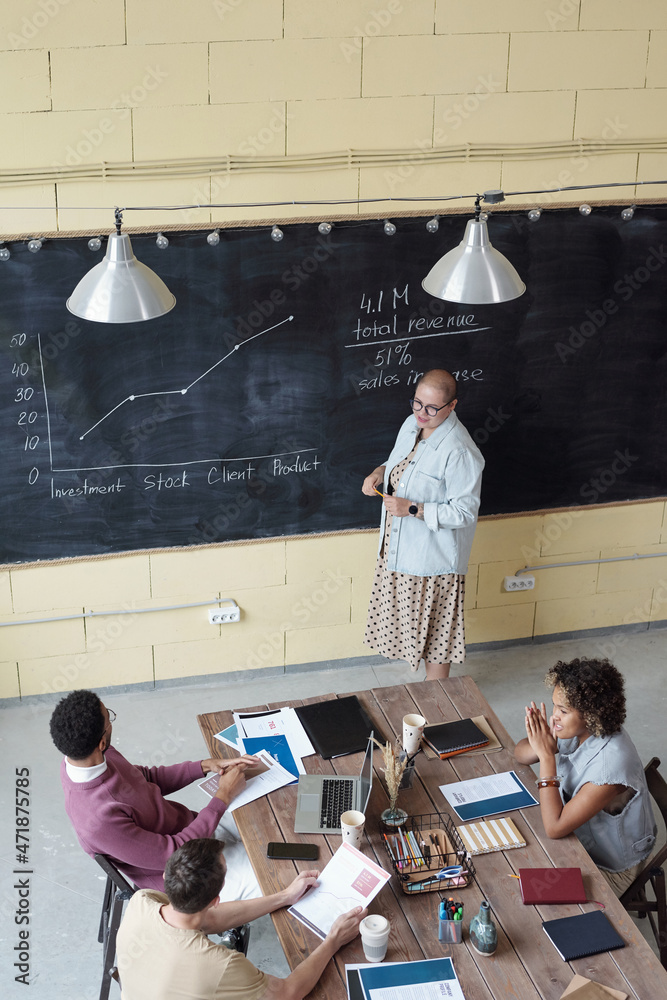 Poster young female coach standing by blackboard with graph and other financial data while explaining it to