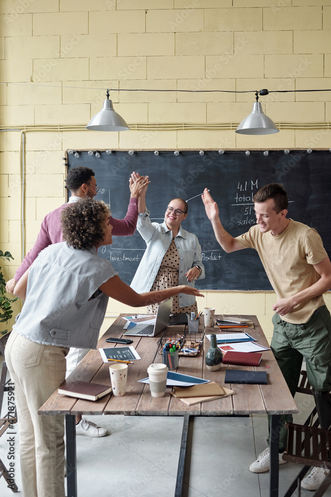 Poster two pairs of young cheerful cow-rkers touching by their palms while standing by table with papers, d