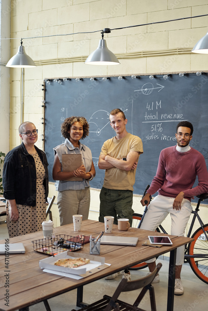Poster happy young economists standing by table with drinks, pizza and gadgets in large openspace office an