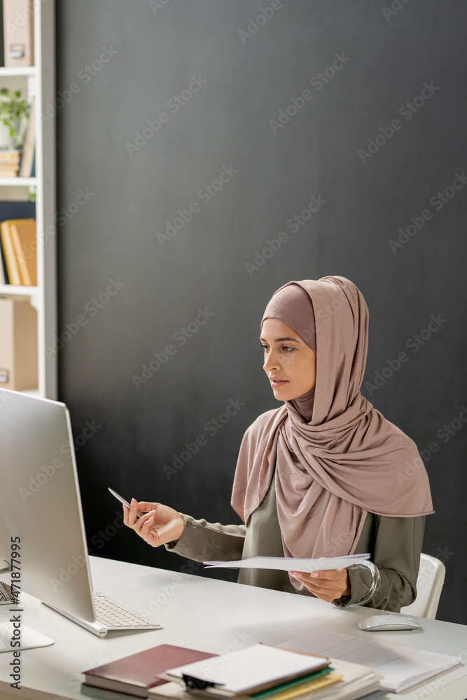 Poster young female teacher in hijab sitting in front of computer and explaining subject of lesson to onlin