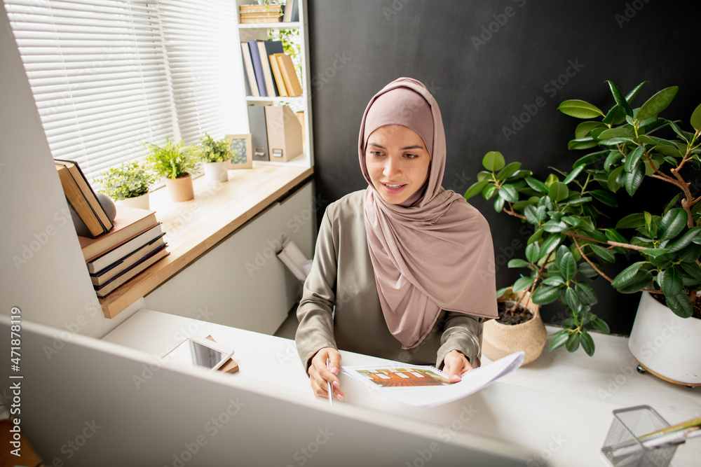 Poster successful young teacher in hijab looking at computer screen during online lesson while answering qu