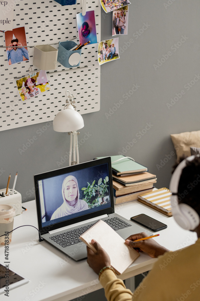 Poster African guy with pencil and notepad sitting by desk in front of laptop and looking at teacher in hijab explaining lesson points