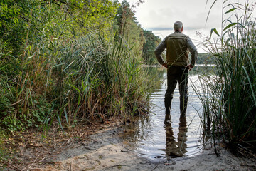 A man stands in the water with his rubber boots and looks out over the lake admiring the beauty of...