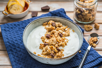 Breakfast or dessert, unsweetened yogurt with granola, dark chocolate, hazelnuts and sliced banana in a ceramic bowl on a wooden background. Breakfast recipes.