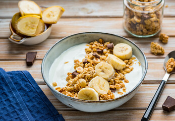 Breakfast or dessert, unsweetened yogurt with granola, dark chocolate, hazelnuts and sliced banana in a ceramic bowl on a wooden background. Breakfast recipes.