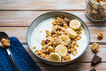 Breakfast or dessert, unsweetened yogurt with granola, dark chocolate, hazelnuts and sliced banana in a ceramic bowl on a wooden background. Breakfast recipes.