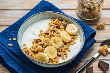 Breakfast or dessert, unsweetened yogurt with granola, dark chocolate, hazelnuts and sliced banana in a ceramic bowl on a wooden background. Breakfast recipes.
