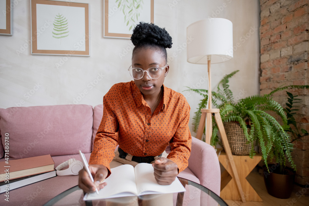 Poster young african woman in eyeglasses making notes in copybook while sitting by table in front of comput