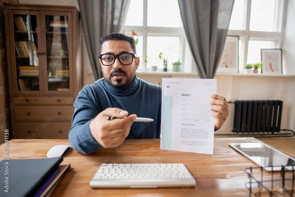 Poster Confident teacher of economics pointing at financial document with budget graph while sitting in front of computer