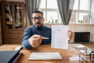 Confident teacher of economics pointing at financial document with budget graph while sitting in front of computer