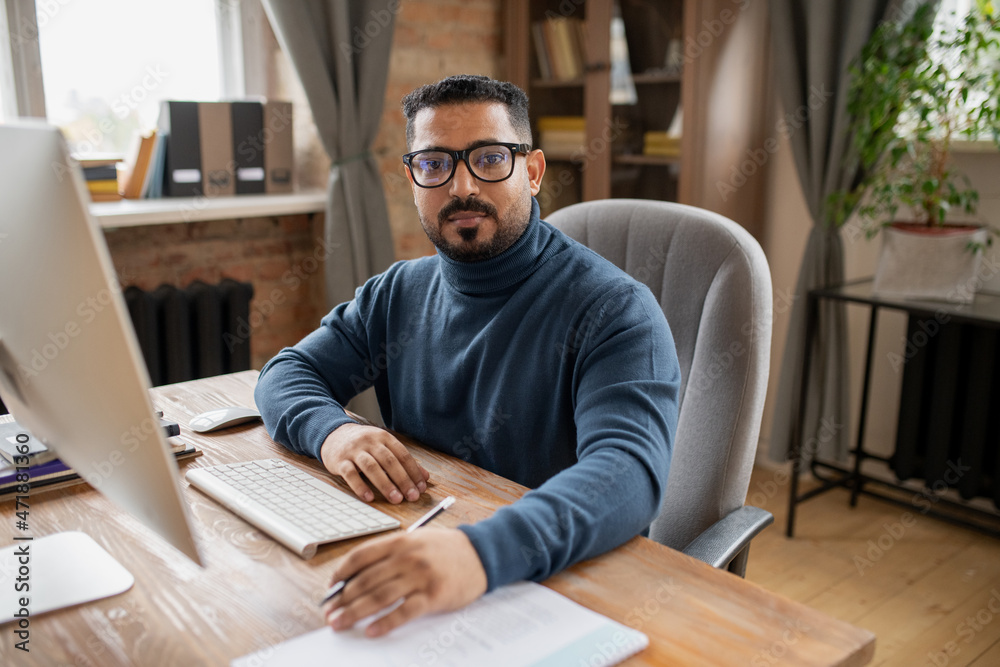 Poster serious middle aged teacher looking at camera while sitting by desk in front of computer monitor in 
