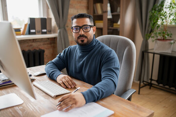 Obraz premium Serious middle aged teacher looking at camera while sitting by desk in front of computer monitor in home environment