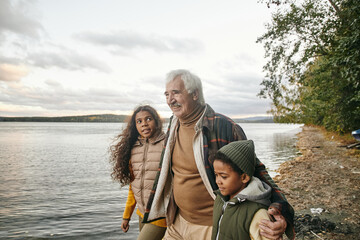 Cute biracial girl looking at her grandfather and listening to him during walk along waterside on windy autumn day
