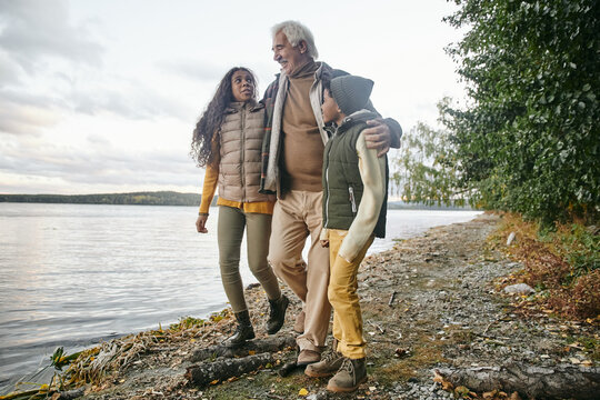 Happy senior man embracing his two interracial grandchildren while moving along waterside and chatting to them