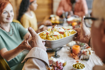 Hand of mature man passing bowl with baked pasta to his wife while both sitting by festive table against their family - obrazy, fototapety, plakaty