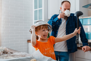 Happy daughter and father playing in kitchen in morning. Child kid girl put colander bowl on her head with ladle in hand she have fun and laughs Plays game. Happy time together family in cooking time