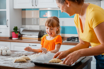 Happy laughing mother and daughter preparing croissants baking in the kitchen together. Family mom and child kid enjoys common leisure time cooking food in kitchen