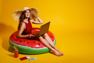 Happy girl in straw hat sits in inflatable watermelon ring uses pc on yellow background. traveler. 