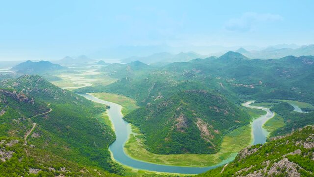 Blue Crnojevica river flowing curves through green valley toward distant mountains of lake Skadar in Montenegro.