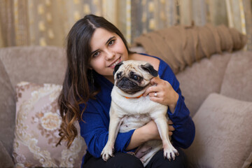 Young beautiful girl sits on a soft sofa with her beloved pug dog