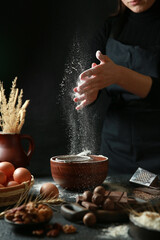 The culinary process. Flour is poured through a sieve into a deep ceramic bowl. Cook in an apron with hands. Chocolate, nuts, eggs on a black table. Background image, copy space