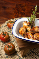 Top view of white tray with roasted chicken wings with rosemary, on wooden table with tomatoes and mat, vertical, with copy space