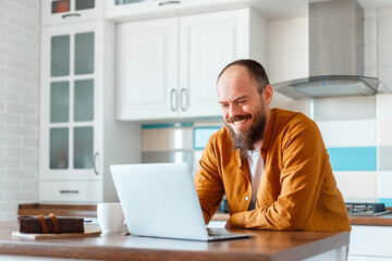 Young man working using laptop in kitchen in home interior. Smiling Freelancer works remotely from home. Happy man owner is doing paperwork using laptop indoors. Bald Caucasian 30s