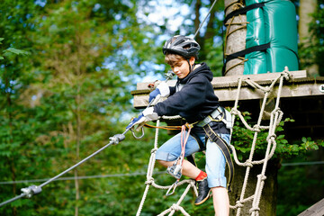 School boy in forest adventure park. Acitve child, kid in helmet climbs on high rope trail. Agility skills and climbing outdoor amusement center for children. Outdoors activity for kid and families.