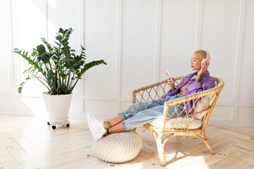 Fototapeta na wymiar Relaxed woman wearing wireless headphones and using smartphone, sitting in wicker chair at home, free space