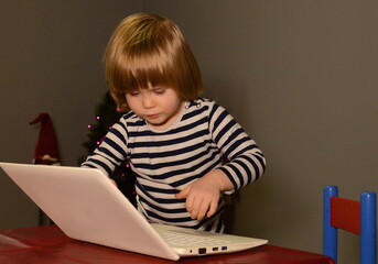 A three-year-old boy is writing a letter to Santa Claus, sitting at a laptop. against the background of the Christmas tree. Christmas concept. close-up. Gray background.