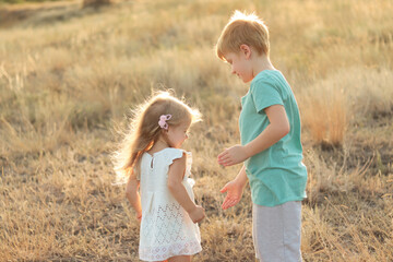 Boy and girl play together in the park. The children laugh. Brother and sister walking in a nature park among thick grass. The girl is wearing a white dress. A boy in a green T-shirt and gray shorts.