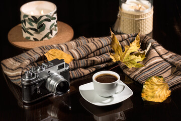 Autumn coffee scene eith vintage camera. A cup of coffee and vintage photo camera on a dark glass table and a warm scarf on a background of autumn leaves. Still life concept with coffee and old camera