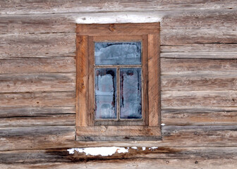A window in a wooden frame is installed in the wall of a log cabin. Frosty patterns formed on the window glass. There is some snow on the log of the log house under the window.