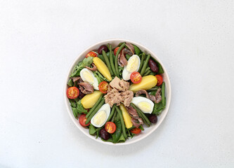 Nicoise salad with tomatoes, eggs, tuna, olives, green beans and anchovies on a white plate, isolated, central composition, top view