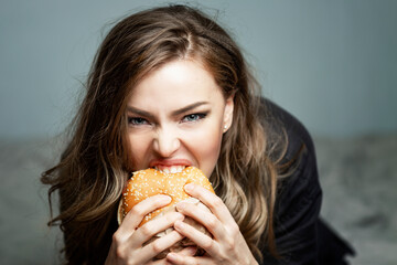 Hungry young woman eating a hamburger. Beautiful blonde. Junk food and quick bites. Gray background. Close-up.