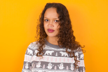 Portrait of dissatisfied teenager girl wearing winter knitted sweater against yellow background...