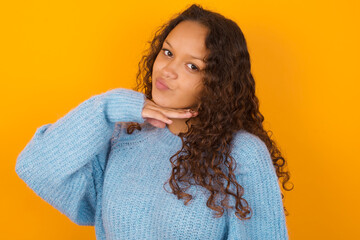 Teenager girl wearing blue sweater over yellow background cutting throat with hand as knife,...
