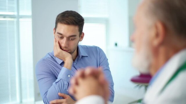 Close-up view from back of mature gray-haired doctor to sad male patient explaining problem sitting at desk during consultation at hospital. Senior physician telling bad news to unhappy young man.