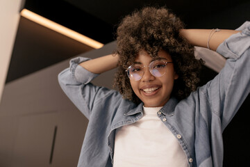 close-up of beautiful young african person in transparent glasses on isolated background. Brunette with short curly hair in light casual outfit holds hands on her head. People and free time concept