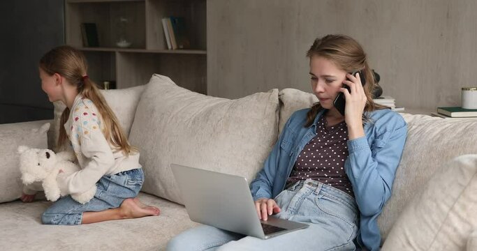 Caring young freelance mom engaged in multitasking discuss affairs on phone before pc tickle cute preteen daughter playing with toy on sofa. Businesswoman combine upbringing child and doing business