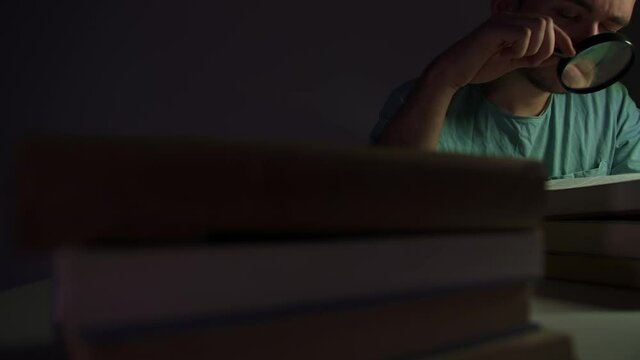 A young guy sits at a table in low light, and reads a book, he is preparing for an exam, and memorizes material, he is dressed in a blue t-shirt, there is no light in the room