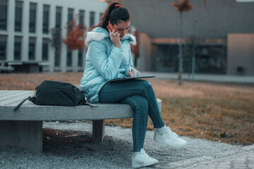 Chica joven de pelo castaño con mascarilla llamando por su telefono movil mientras revisa los apuntes de la universidad para estudiar	