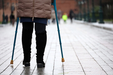 Woman walking with crutches on a city street on people background, female legs on sidewalk. Concept of disability, limping person with injury - obrazy, fototapety, plakaty