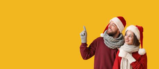 Happy young couple in stylish winter clothes and Santa Claus hats showing something on color background with space for text
