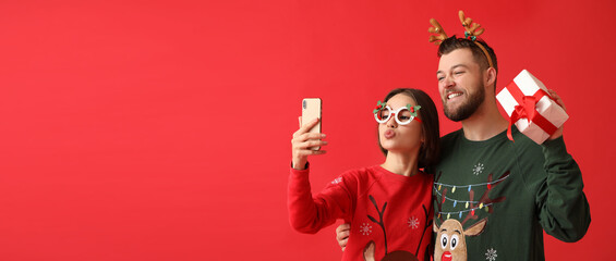 Happy young couple in stylish Christmas clothes and with gift taking selfie on color background...